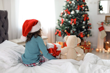 Obraz na płótnie Canvas Cute little child in Santa hat with toy bear and Christmas gift box sitting on bed at home