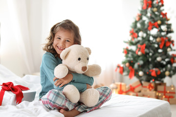 Cute little child with toy bear and Christmas gift box sitting on bed at home