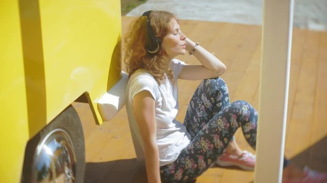 young woman outdoors by yellow vagon car listening music in headphones using smartphone - relaxing, enjoying, concept of technology and travel