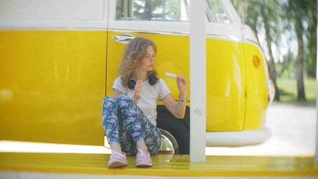 young woman outdoors by yellow vagon car listening music in headphones using smartphone - relaxing, enjoying, concept of technology and travel