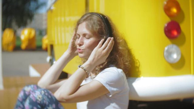 young woman outdoors by yellow vagon car listening music in headphones using smartphone - relaxing, enjoying, concept of technology and travel