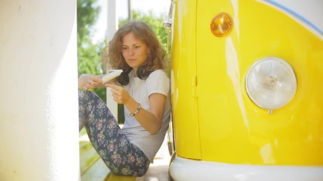 young woman outdoors by yellow vagon car listening music in headphones using smartphone - relaxing, enjoying, concept of technology and travel