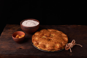 On a dark wooden table, fashionable bakeries, a bowl with flour, apricot cake and tools stand on table