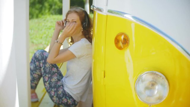 young woman outdoors by yellow vagon car listening music in headphones using smartphone - relaxing, enjoying, concept of technology and travel