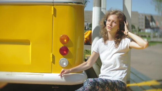 young woman outdoors by yellow vagon car listening music in headphones using smartphone - relaxing, enjoying, concept of technology and travel