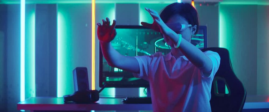 East Asian Pro Gamer Wearing Virtual Reality Headset Plays in a Online Video Game, Gesturing. Gameplay Shown on Computer Monitor. Cool Retro Neon Colors in the Room. Shot with Anamorphic Lens.