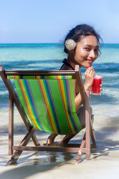 Big head girl with a white flower in her hair relaxed by the sea with red juice. Beautiful woman sitting in a deck chair on the sea beach. 