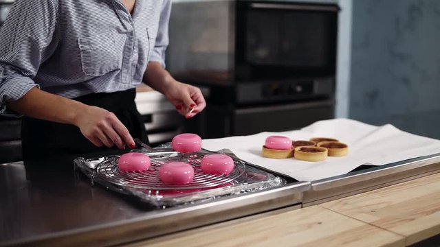 Confectioner woman working on modern kitchen. Preparing modern, french desserts - mousse with pink mirror glaze putting on top baked tartles. Close up footage