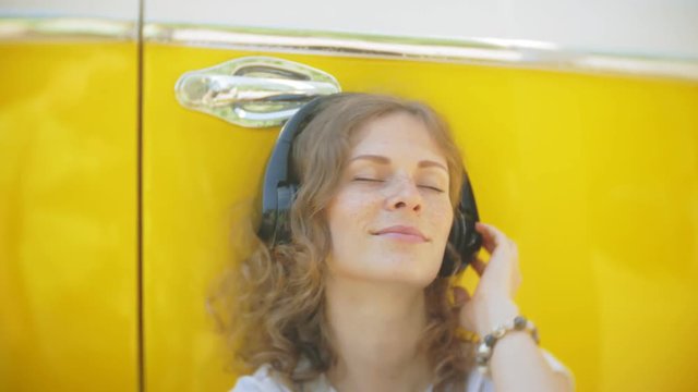 young woman outdoors by yellow vagon car listening music in headphones using smartphone - relaxing, enjoying, concept of technology and travel