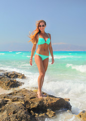 Fototapeta premium Athletic woman in cyan blue bikini standing on rocks with turquoise sea behind her
