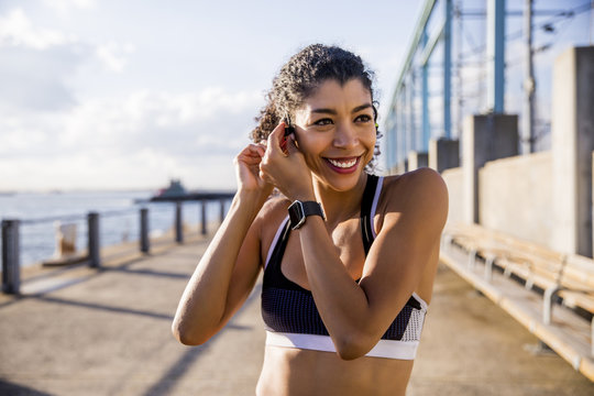 Smiling female athlete adjusting headset while standing on footpath against sky