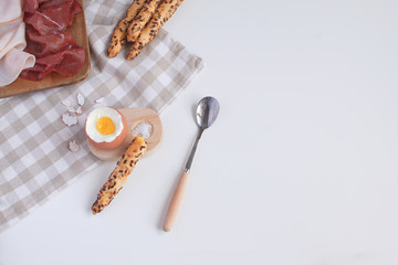Served breakfast boiled egg in wooden egg cup with breadsticks ham salt spoon and checkered napkin White background Top view Minimalism Copy space