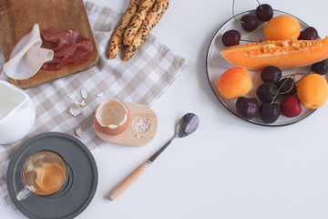 Perfectly served breakfast boiled egg in wooden egg cup with breadsticks ham melon cherry apricots coffee salt spoon and checkered napkin White background Top view Copy space