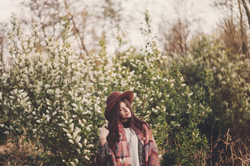 stylish hipster girl in hat and poncho with beautiful hair standing in park in evening sunshine....