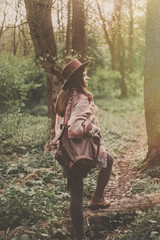 stylish hipster traveler girl in hat with backpack walking in woods in evening sunshine. bohemian fashionable woman exploring in sunlight. space for text. atmospheric moment. wanderlust