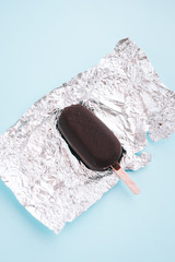 Dark chocolate ice-cream, popsicle on a silver paper wrap from above. Pastel blue background....