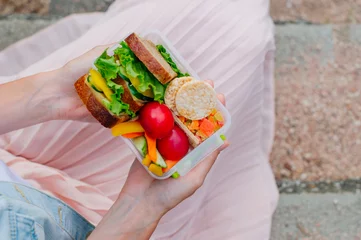 Gordijnen Close up of young girl holding lunch box filled with sandwich, crispbreads, fruits and vegetables © lithiumphoto