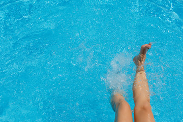 Girl is swinging legs in a turquoise pool creating splashes in the sunny day