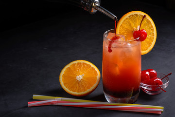 Making Tequila Sunrise Cocktail. Grenadine slowly poured into a glass of ice, orange juice and tequila. Dark stone background with space for your text.