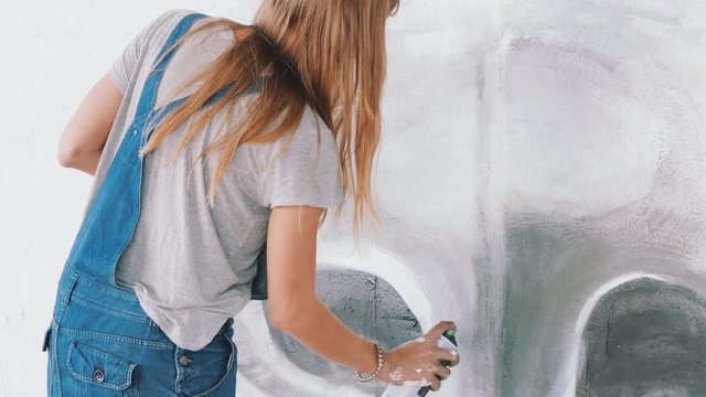 Beautiful Young Girl making graffiti with aerosol spray on urban street wall. Cinematic toned footage. Creative art. Talented student in denim overalls drawing picture.