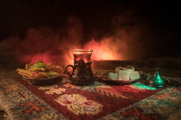 Arabian tea in glass with eastern snacks on a carpet on dark background with lights and smoke. Eastern tea concept. Empty space.