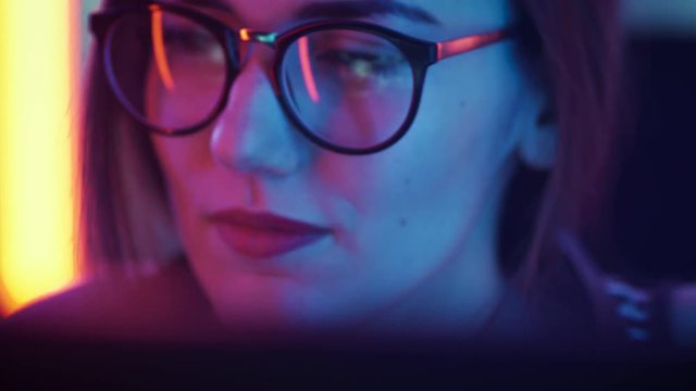 Portrait of the Beautiful Young Girl Sitting Before Computer, Browsing in Internet, Playing Online Games, Streaming. Cute Girls Wearing Glasses in the Cool Retro Neon Lit Room.