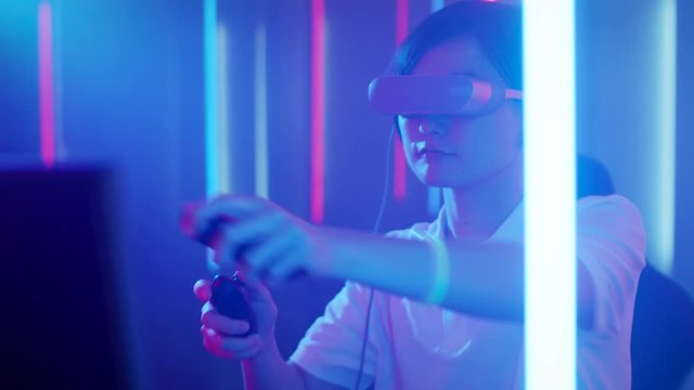 East Asian Pro Gamer Puts On Virtual Reality Headset Plays Online Video Game with Joysticks / Controllers. Cool Retro Neon Colors in the Room.