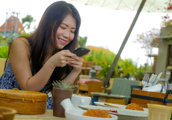 young beautiful and happy Asian Chinese woman using internet social media app on mobile phone smiling cheerful enjoying breakfast relaxed outdoors