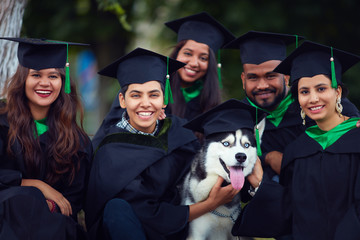 portrait of happy indian students friends in graduation gowns with pet dog