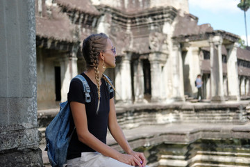 Fototapeta na wymiar Young sun-tanned woman tourist with pigtails sitting near ancient temple. Man silhouette in background. Angkor Wat, Cambodia