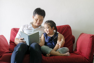 Father and daughter are reading books.Sitting on a red sofa.