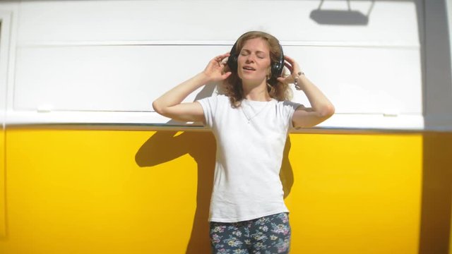young woman outdoors by yellow vagon car listening music in headphones using smartphone - relaxing, enjoying, concept of technology and travel
