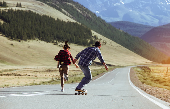 Happy young couple having fun with longboard on the road. Young man and woman skating together on a sunny day in mountain. - Powered by Adobe