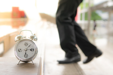 Blured legs with plants and black leather shoes are walking up the steps behind alarm clock in the morning