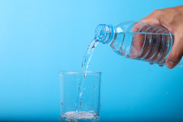 Closeup of pouring mineral water from bottle into glass by hand with blue background. 