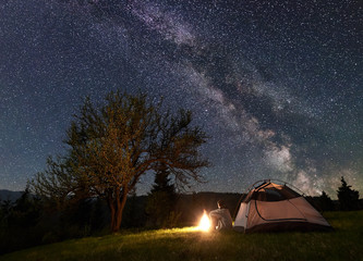 Fototapeta na wymiar Male backpacker sitting alone in front of tourist tent at campfire on grassy valley enjoying night blue starry sky with Milky way. Beauty of nature, tourism and traveling concept. Mountain camping