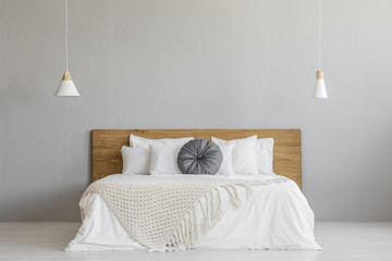 Knit blanket on wooden bed against grey wall in minimal bedroom interior with lamps. Real photo - obrazy, fototapety, plakaty