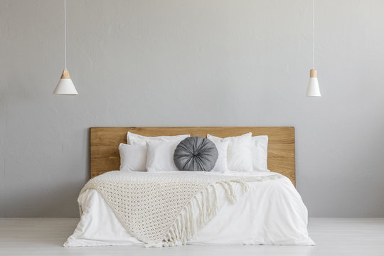 Knit blanket on wooden bed against grey wall in minimal bedroom interior with lamps. Real photo