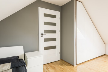 White cabinet between white door and bed with sheets in grey bedroom interior on attic. Real photo - obrazy, fototapety, plakaty