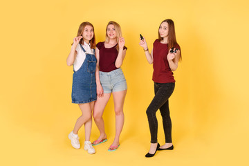Fototapeta premium three young girls on a yellow isolated background look directly into the camera. two girls stretch a chewing gum, a third holds the bottles with nail Polish