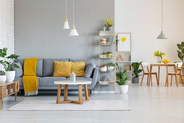 Lamps above wooden table in open space interior with yellow blanket on grey sofa. Real photo