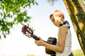 Young boy out of focus plays the guitar standing under the tree and enjoying a beautiful summer day in the park Handsome kid relaxing outdoor playing a musical instrument with straw hat and bandana
