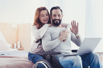 Video call. Cheerful young woman hugging her disabled husband while having a pleasant video call