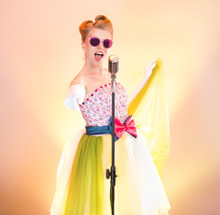 Happy girl singer in pink glasses smiles, have fun, sings into retro microphone, points shows by hand finger into camera. Stylish teenager musician in funny retro dress on colored rainbow background