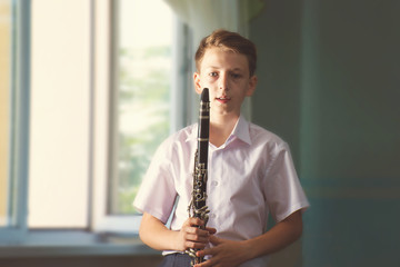 The boy stands near the window and holds a black clarinet in his hands, looks into the camera. Musicology, music education and education.
