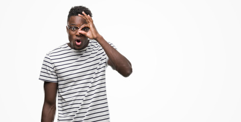 Young african american man wearing glasses and navy t-shirt doing ok gesture shocked with surprised face, eye looking through fingers. Unbelieving expression.
