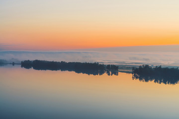 Broad mystical river flows along diagonal shore with silhouette of trees and thick fog. Gold glow in predawn sky. Calm morning atmospheric landscape of majestic nature in warm tones.