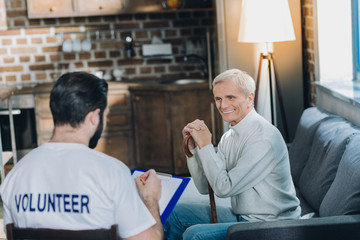 Valuable answers. Well-built dark-haired volunteer talking with an aged man and conducting a poll while sitting on the chair and the old man smiling