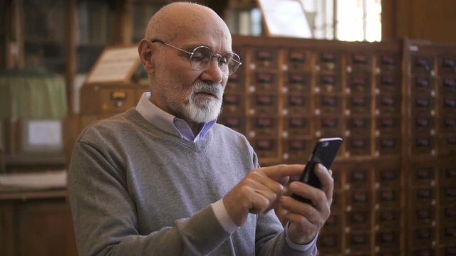 Respectable professor is using smartphone standing in university library, elderly bearded man is looking at phone screen, typing during working day in reading room. Concept: mature person, lifestyle
