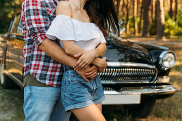 embrace.Love and affection between a young couple at the park, near the old car. a guy in a plaid plane and jeans, a girl in shorts and a white jacket. They get together in the forest for a walk.